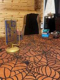 Tarot reading table with gold stand holding a 'Death' tarot card, orange spiderweb tablecloth, stacked card decks and a crystal ball against a wooden-paneled cozy reading room backdrop.