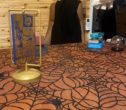 Tarot reading table with gold stand holding a 'Death' tarot card, orange spiderweb tablecloth, stacked card decks and a crystal ball against a wooden-paneled cozy reading room backdrop.