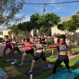 Outdoor yoga class in a sunny suburban backyard with a group of women smiling and practicing Warrior II on mats under string lights and leafy trees