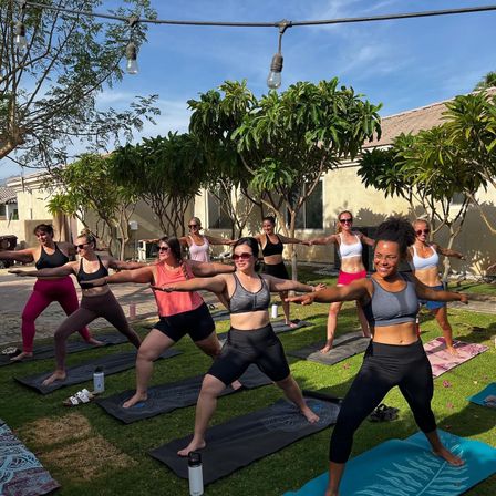 Smiling women in an outdoor yoga class striking Warrior II on mats across a sunny suburban backyard lawn under string lights and leafy trees.