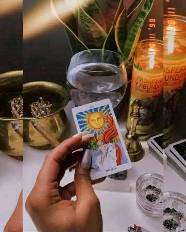 Hand holding The Sun tarot card above a cozy indoor altar for a tarot reading — lit “Prosperidad y Abundancia” candles, glass of water, brass bowl, small ashtray with ashes and a potted snake plant.