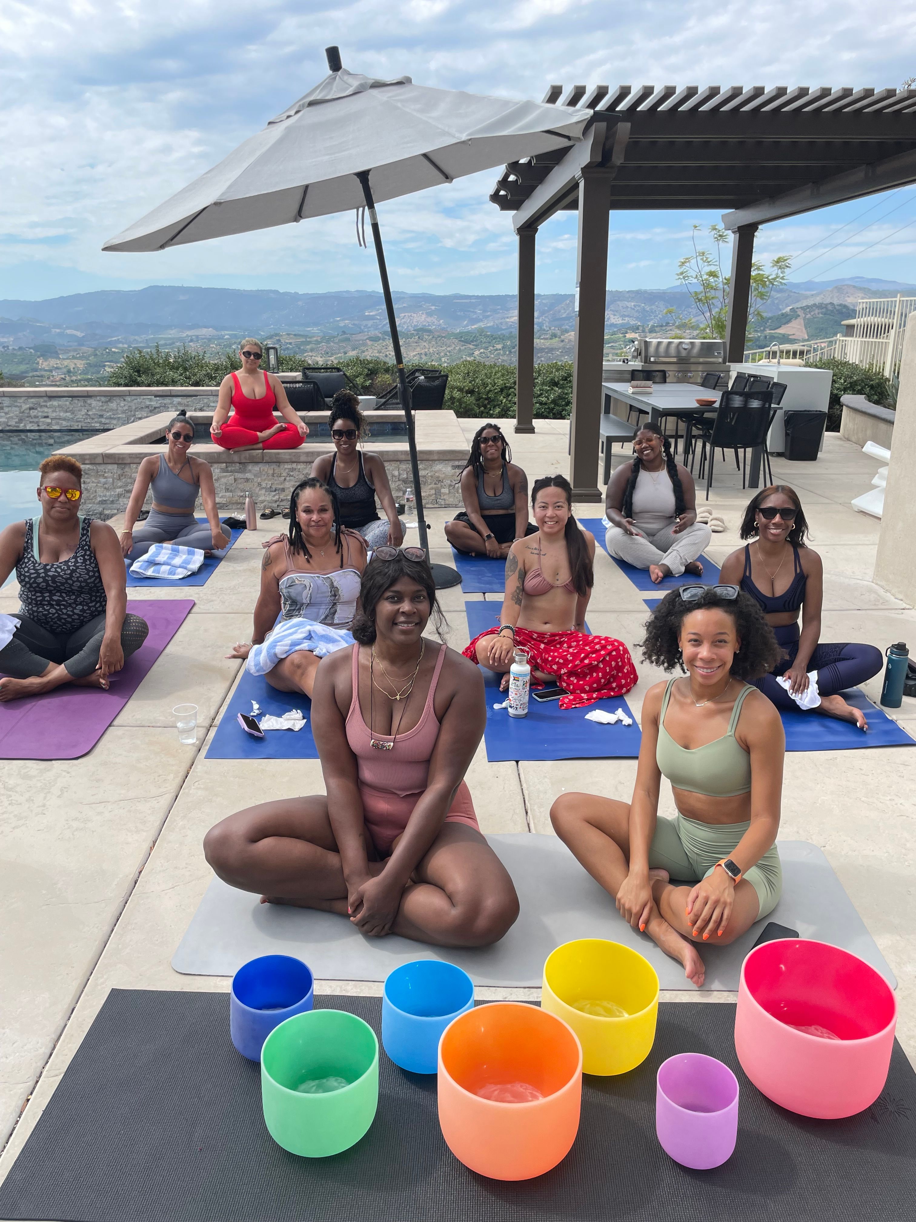 Outdoor poolside yoga and sound-bath session with a group of women seated on mats under an umbrella and pergola, colorful crystal singing bowls in the foreground and rolling hills and mountains in the background.