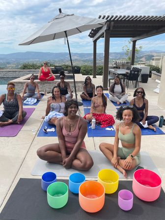 Outdoor poolside yoga and sound-bath session with a group of women seated on mats under an umbrella and pergola, colorful crystal singing bowls in the foreground and rolling hills and mountains in the background.