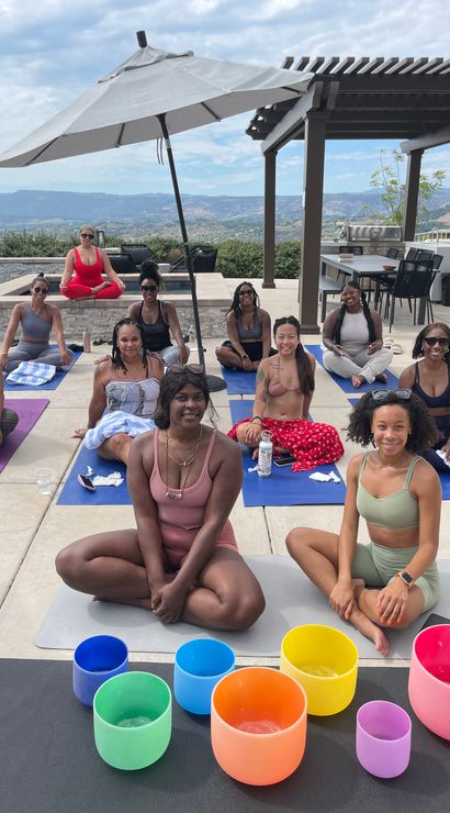 Outdoor poolside yoga and sound-bath session with a group of women seated on mats under an umbrella and pergola, colorful crystal singing bowls in the foreground and rolling hills and mountains in the background.