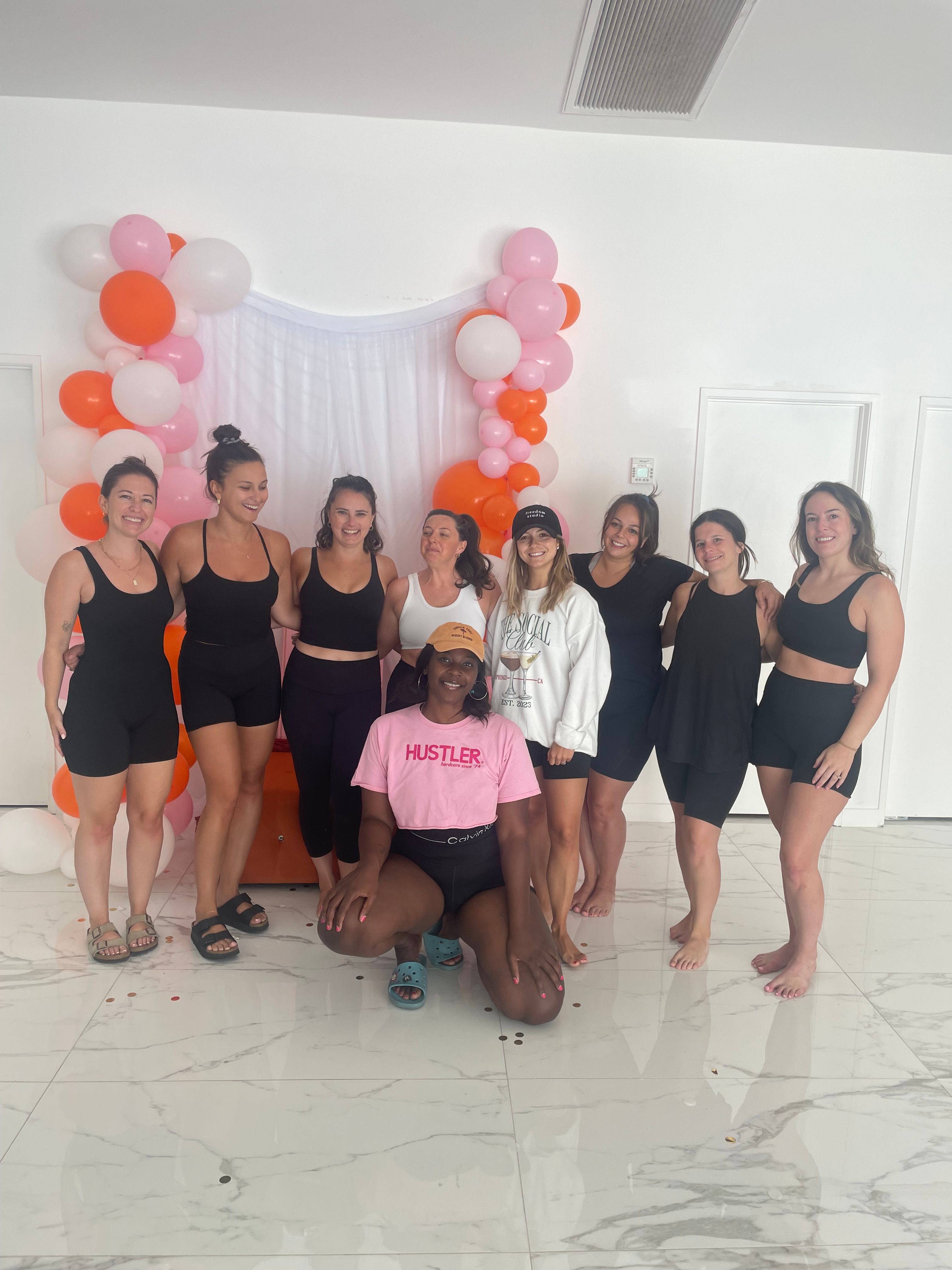 Nine women in activewear smiling and posing in a bright white indoor studio in front of a pink, orange and white balloon arch and sheer backdrop — fun group fitness/wellness class vibe.