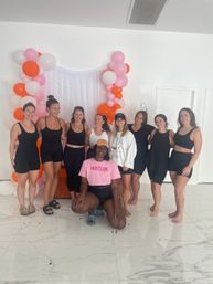Nine women in activewear smiling and posing in a bright white indoor studio in front of a pink, orange and white balloon arch and sheer backdrop — fun group fitness/wellness class vibe.