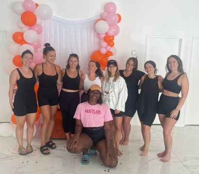 Nine women in activewear smiling and posing in a bright white indoor studio in front of a pink, orange and white balloon arch and sheer backdrop — fun group fitness/wellness class vibe.