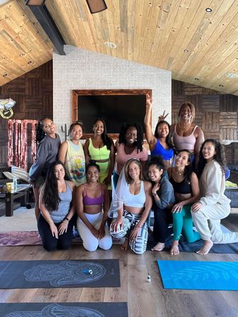 Smiling group of women in colorful activewear posing on yoga mats inside a cozy cabin-style indoor yoga studio with wooden ceiling and patterned wall — upbeat wellness retreat vibe.