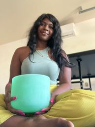 Smiling person seated cross-legged in a home meditation scene, holding a mint-green crystal singing bowl while wearing a ribbed tank top and yellow pants — peaceful wellness moment.