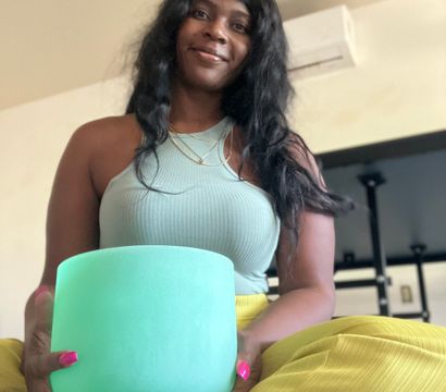 Smiling person seated cross-legged in a home meditation scene, holding a mint-green crystal singing bowl while wearing a ribbed tank top and yellow pants — peaceful wellness moment.