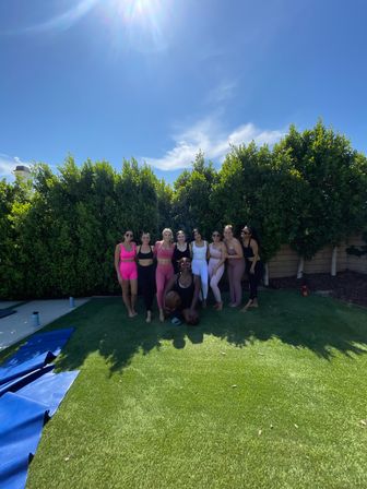 Group fitness scene: smiling women in colorful activewear posing on a sunny suburban backyard lawn with yoga mats, tall hedges and clear blue sky — outdoor workout/backyard yoga session.