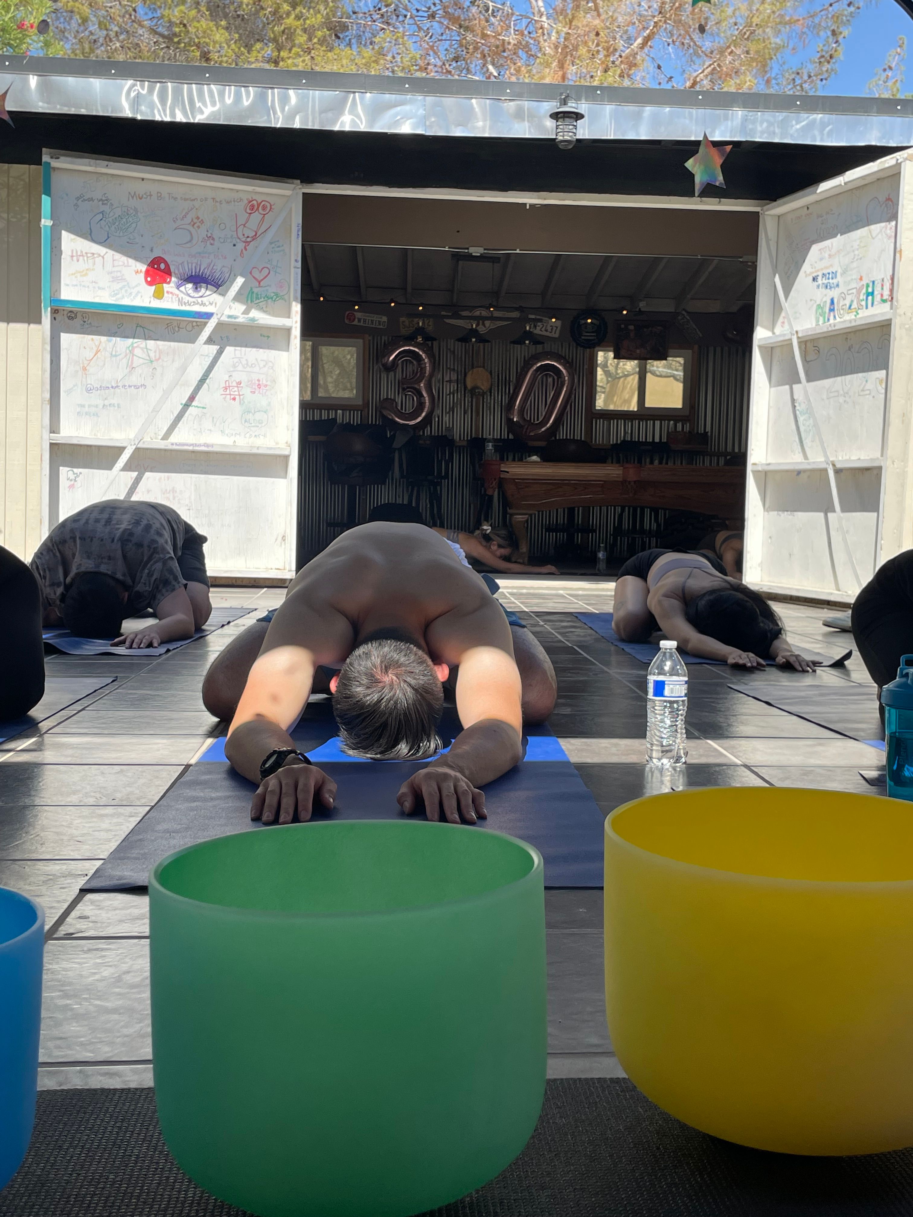 Open-air yoga class with participants in child's pose on mats, colorful sound bowls in the foreground, a water bottle on the floor, and large '30' balloons inside a rustic studio
