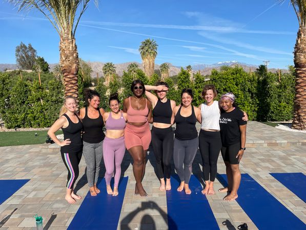 Eight women in colorful activewear smiling and posing barefoot on blue yoga mats on a sunny palm-lined patio with mountains and a clear blue sky — outdoor group yoga session.