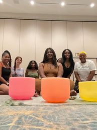 Six women seated on a rug in a large conference room, smiling behind pink, orange and yellow crystal singing bowls during a sound-bath meditation workshop