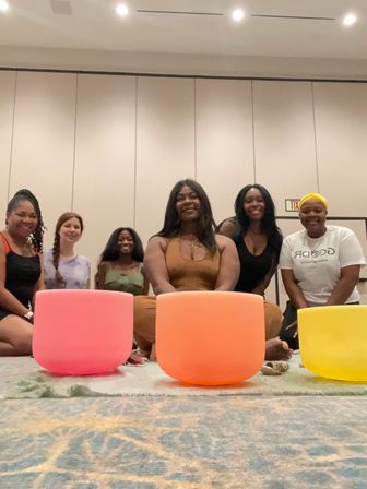 Six women smiling and seated on a carpeted conference room floor behind three colorful crystal singing bowls (pink, orange, yellow) during an indoor sound-bath wellness circle.
