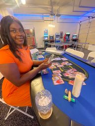 Person in an orange dress smiling while arranging tarot cards on a blue gaming table in a garage-style game room with arcade cabinets, a ping-pong table, a lit candle and a drink with ice.