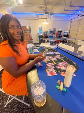 Smiling woman holding tarot cards over a tarot spread on a game-room poker table with candle and drink, LED-lit garage space with arcade cabinets and a ping-pong table in the background.