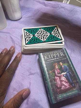Stack of teal-and-white Celtic-back Kipper divination cards and boxed Kipper fortune-telling deck on purple cloth, with a manicured hand and two white candles for a cozy cartomancy setup.