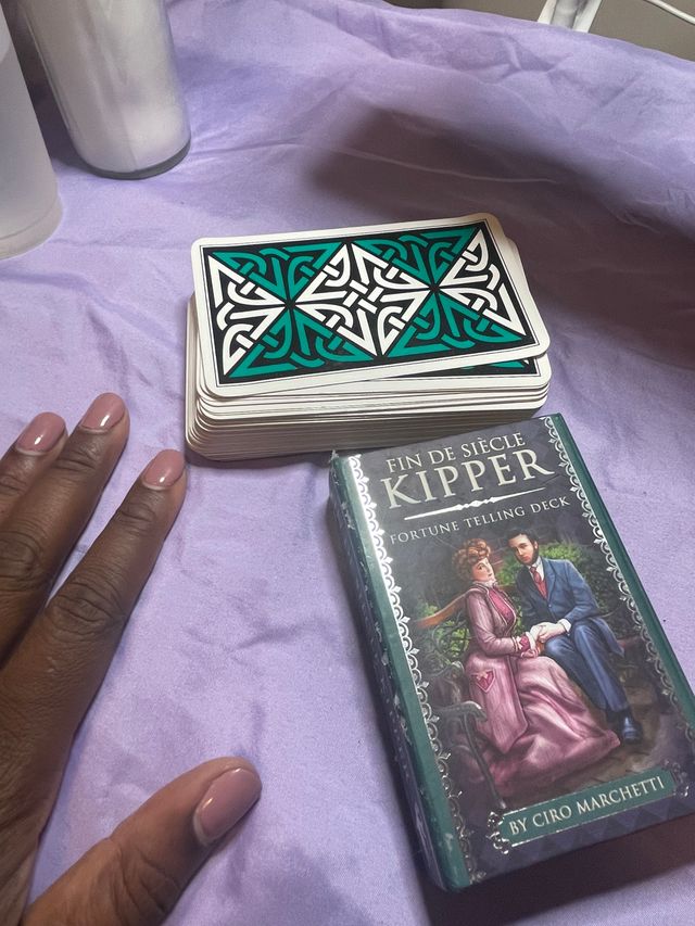 Stack of teal-and-black patterned fortune-telling cards and a Kipper fortune-telling deck box on a lavender sheet, with a hand showing glossy pale pink nails nearby.