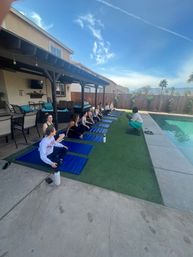 Relaxed outdoor group yoga class on blue mats beside a suburban backyard pool under a patio pergola, instructor leading seated poses with sunny sky and palm trees in the background.