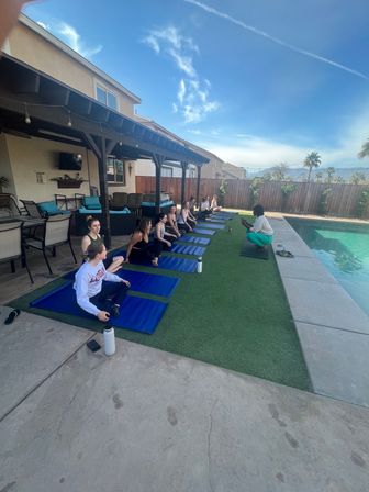 Relaxed outdoor group yoga class on blue mats beside a suburban backyard pool under a patio pergola, instructor leading seated poses with sunny sky and palm trees in the background.