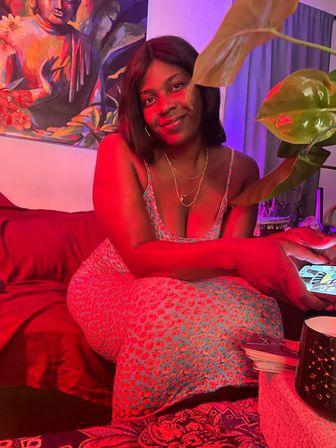 Woman smiling on a cozy couch bathed in red and purple ambient light, wearing a floral dress and holding a smartphone, with a leafy plant, Buddha painting, mug and a small deck of cards nearby.