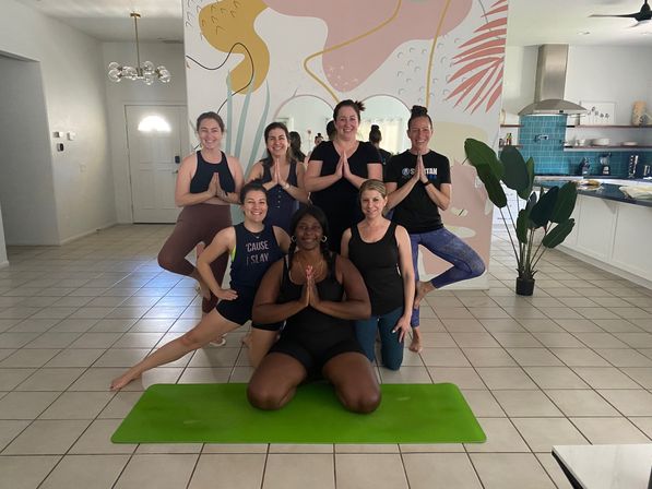 Smiling group yoga class indoors on tiled floor with colorful abstract mural and a green yoga mat, participants posing with hands in prayer.