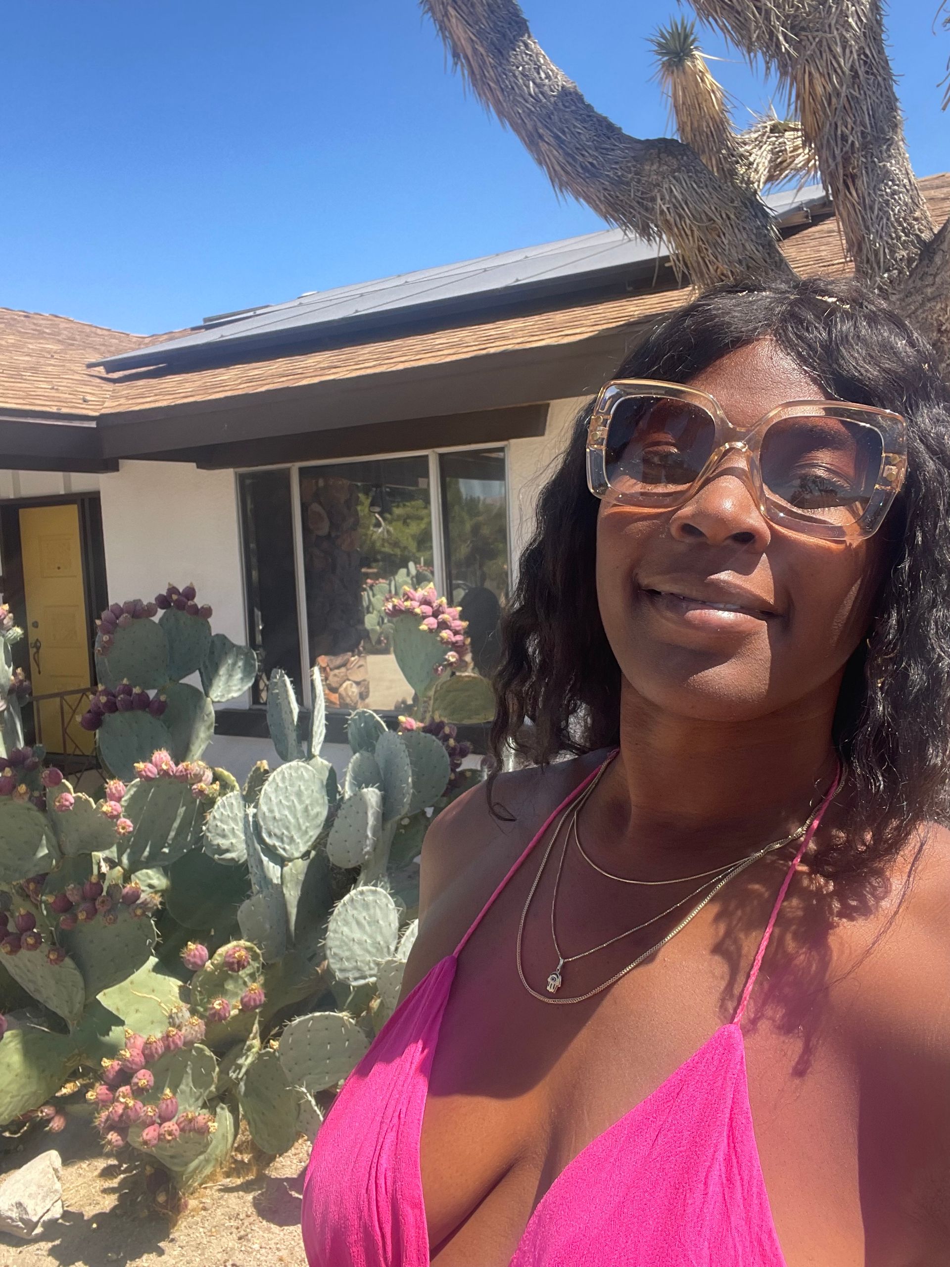 Sun-soaked selfie of a woman in a bright pink halter dress and oversized sunglasses in a southwestern desert front yard with blooming prickly pear cactus, a low stucco house with solar panels, and clear blue sky.