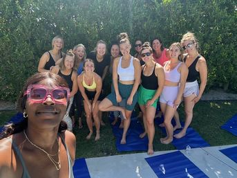 Women posing for a selfie at an outdoor backyard yoga/fitness class on blue mats, wearing colorful athletic wear and sunglasses, barefoot on grass in front of a tall green hedge on a sunny day; woman with pink sunglasses holds the camera.