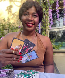 Smiling woman at a garden patio doing an outdoor tarot reading, holding three colorful tarot cards with red nails and rings, purple hanging flowers and a tarot deck on the table.