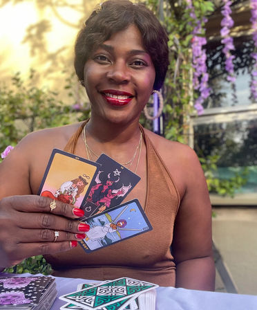 Smiling woman at a garden patio doing an outdoor tarot reading, holding three colorful tarot cards with red nails and rings, purple hanging flowers and a tarot deck on the table.