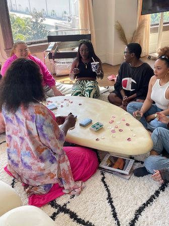 Group of friends seated on a shag rug around a low table in a sunlit loft-style living room, playing cards with scattered rose petals and sipping drinks — cozy weekend gathering.
