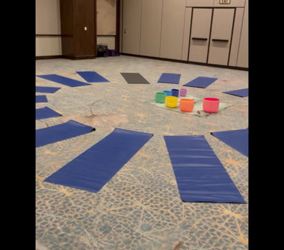 Blue yoga mats arranged in a circle on a patterned carpet inside a conference room surrounding bright, colorful crystal singing bowls for a sound-healing yoga session