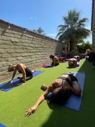 Sunny backyard yoga session with a group stretching on blue mats on green artificial turf beside a beige brick wall and tall palm tree under a clear blue sky.