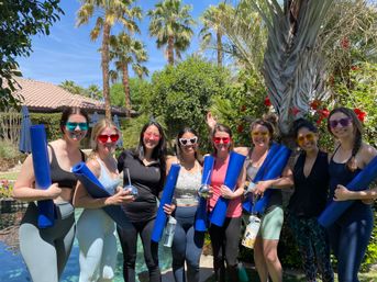 Group of eight people posing poolside in a sunny palm-tree backyard, each holding blue yoga mats and wearing colorful sunglasses for an outdoor yoga/fitness session