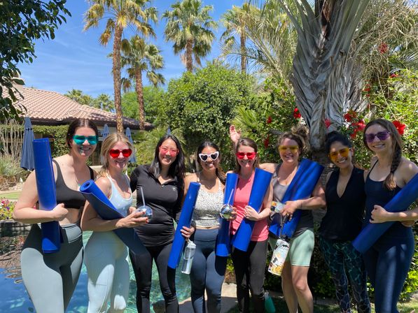 Group of eight people posing poolside in a sunny palm-tree backyard, each holding blue yoga mats and wearing colorful sunglasses for an outdoor yoga/fitness session