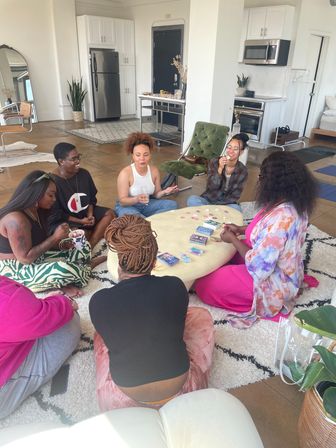 Bright modern loft living room with six women seated on a rug around a low table, enjoying drinks, snacks and a card game during a cozy game night; open kitchen, plants and casual seating in the background.