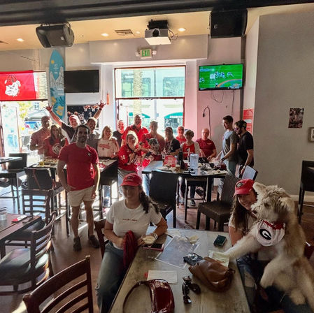 Group of Georgia Bulldogs fans in red gear cheering at a lively downtown sports bar watch party, drinks on tables and a dog wearing a team bandana