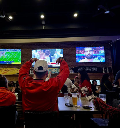 Crowded sports bar interior with a fan in a red jacket and white cap cheering toward wall-mounted TVs showing football and an MMA fight, beers and phone on the table, brick-wall backdrop.