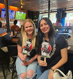 Two friends smiling at a lively sports bar, each holding a bulldog mascot cutout on a stick with TVs showing games and a wooden bar with taps in the background.