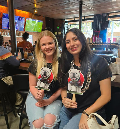 Two friends smiling at a lively sports bar, each holding a bulldog mascot cutout on a stick with TVs showing games and a wooden bar with taps in the background.