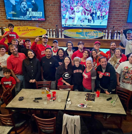 Large group of Georgia Bulldogs fans at a college football watch party in a lively sports bar, wearing red and black team gear, TVs showing the game and surfboard décor on a brick wall