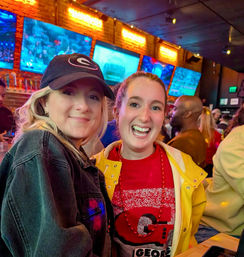 Two smiling women posing in a lively sports bar — one in a black cap and denim jacket, the other in a yellow jacket and red Georgia team shirt with bead necklace, with multiple TVs showing a game and a brick wall behind them.