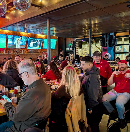Lively crowded sports bar interior with patrons at a long bar, multiple TVs showing games, disco balls and colorful party lighting.