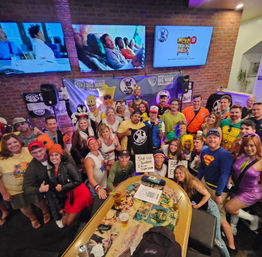 Large group in colorful costumes posing and smiling around a high table with drinks inside a lively brick‑walled bar with multiple TV screens