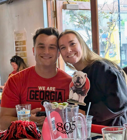 Smiling couple in red "We Are Georgia" shirt posing with a Georgia Bulldogs bulldog cutout at a lively game-day bar table with a pitcher of drinks and fan pom-pom