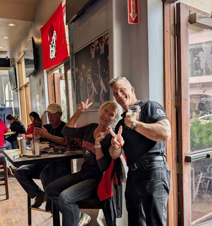 Smiling middle-aged friends flash peace signs inside a sunlit sports bar by an open front door, holding drinks at a high table with other patrons and a red college sports flag on the wall.