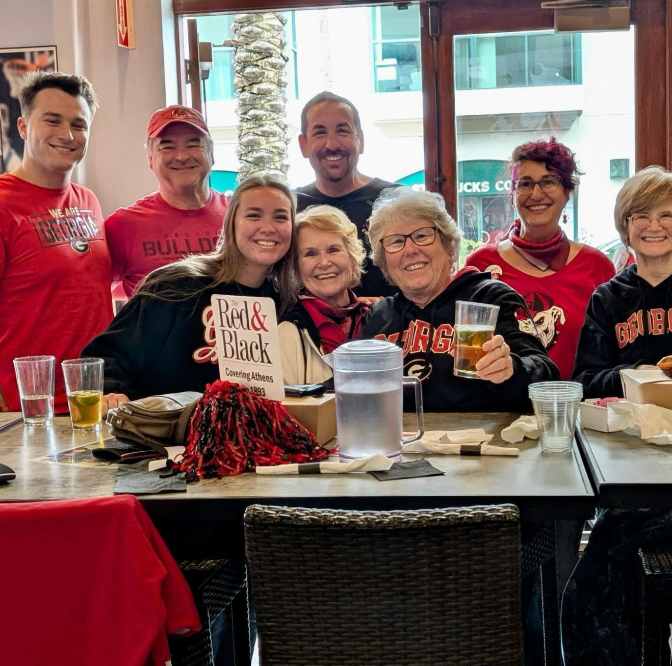 Smiling group of University of Georgia fans in red-and-black UGA gear enjoying drinks at a sports-bar table with a pom-pom — game-day vibe in Athens, GA.