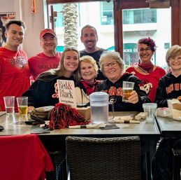 Smiling group of University of Georgia fans in red-and-black UGA gear enjoying drinks at a sports-bar table with a pom-pom — game-day vibe in Athens, GA.