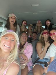 Group selfie of smiling young women in a passenger van wearing summer outfits, baseball caps and pink sunglasses, packed in for a fun road-trip or beach day.